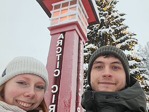 Selbstporträt zweier Studierender, dahinter eine Säule, die den Polarkreis markiert.