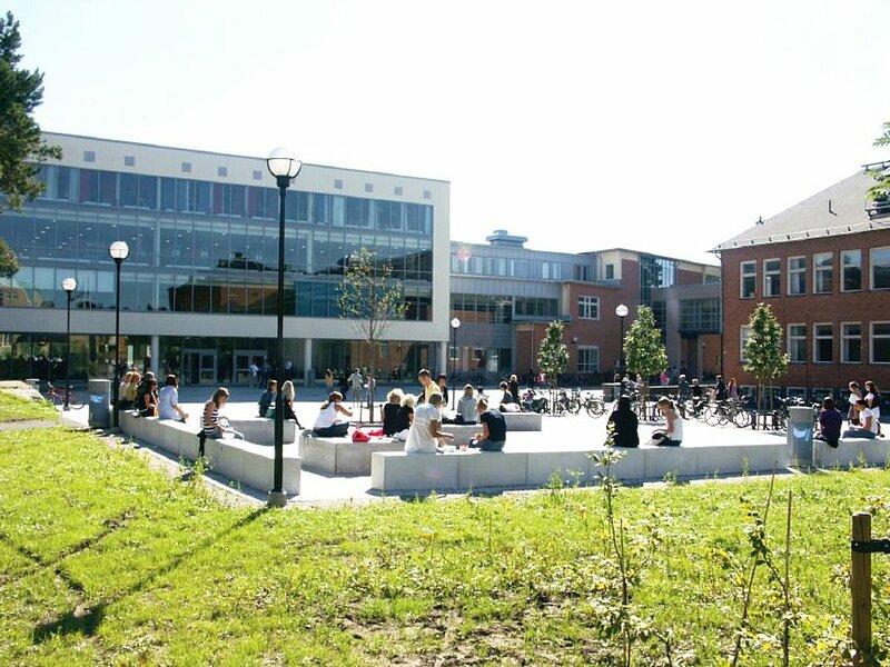 Blick auf Campus im Sommer, Personen auf Steinbänken sitzend lesen oder unterhalten sich.