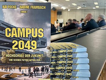 Die Anthologie von vorne zu sehen, dahinter einige Exemplare gestapelt auf einem Tisch im Hörsaal. Dahinter sieht man das lauschende Plenum während der Buchpräsentation. 