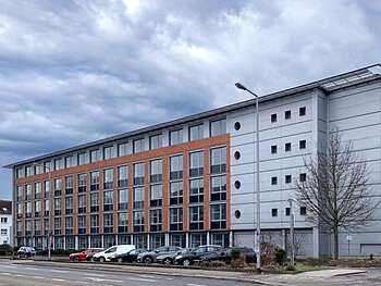 Blick von der Straßenseite auf ein 4-stöckiges Bürogebäude in grau und Terracotta, davor parken 8 Autos.