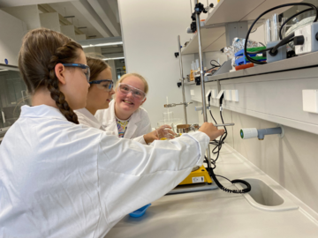 3 Mädchen mit weißem Kittel und Schutzbrillen im Chemie-Labor, 2 davon arbeiten mit einer Waage, eine lacht in die Kamera.