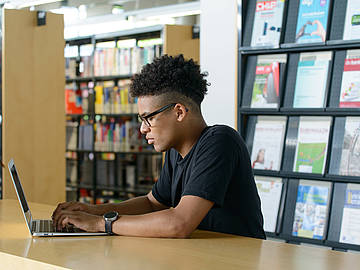 Von der Seite zu sehen: Student mit dunkler Haut, Brille und dunklen Haaren am Laptop in der DHBW-Mannheim-Bibliothek sitzend.