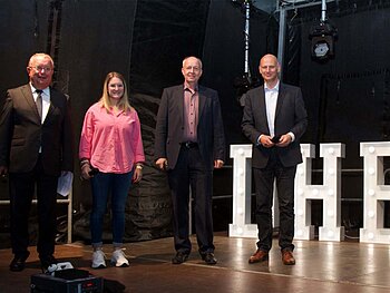 Drei Männer und eine junge Frau stehen in Abendkleidung auf eine Bühne vor Leuchtbuchstaben