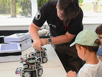 Ein Mitglied der TIGERs zeigt drei Kindern kleine Fußballroboter, die auf dem Tisch stehen 