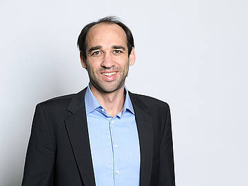 Porträt Prof. Dr. Markus Schief: Dunkle Haare, freundliches Gesicht, Mitte/Ende Dreißig, in schwarzem Anzug und hellblauem Hemd.