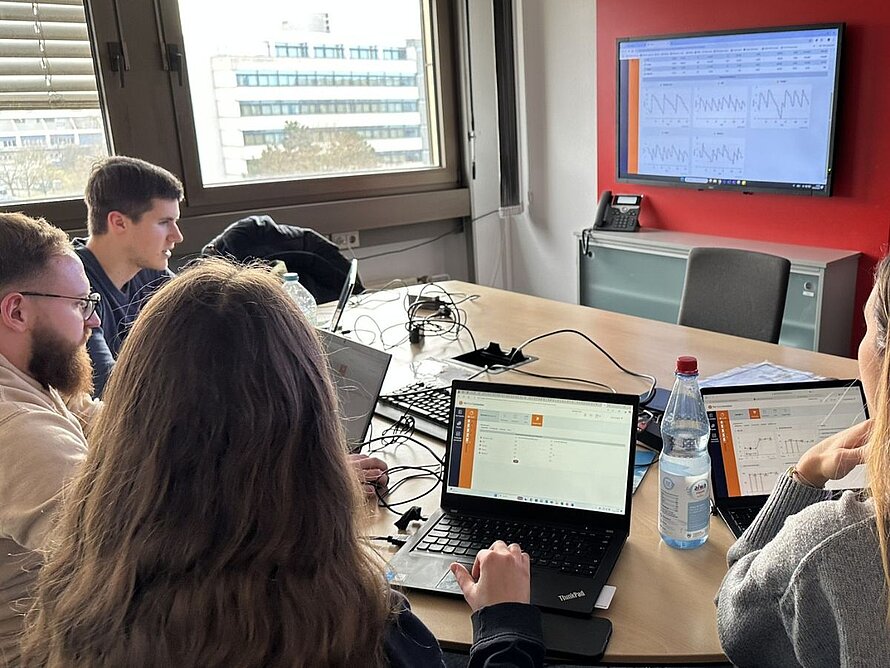 Studierende arbeiten gemeinsam an Laptops in einem modernen Arbeitsraum; im Hintergrund ein Monitor mit Daten-Dashboards.