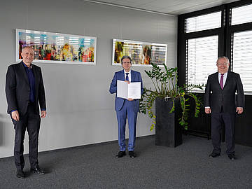 3 Männer im Anzug in einem Konferenzsaal mit der Urkunde zum Dienstjubiläum