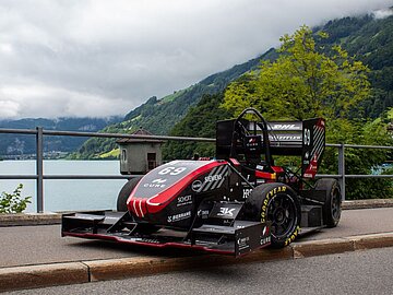 Rennwagen auf einer Brücke mit See und Bergpanorama im Hintergrund. 