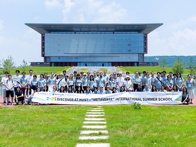 Gruppenbild mit 50 Personen auf einer Wiese, die ein Banner zur Summer School halten. Im Hintergrund ein modernes, verglastes Gebäude. 