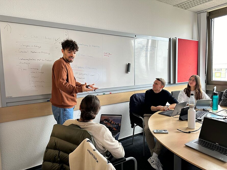 Ein Student präsentiert einer Gruppe an einem runden Tisch Ergebnisse auf einem Whiteboard im Besprechungsraum.
