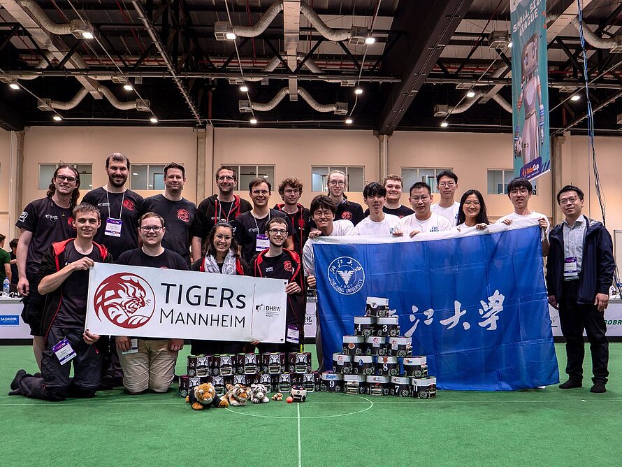 Zwei Mannschaften nebeneinander, links die TIGERs, rechts ZJUNGlict, beide mit ihren Flaggen und den vor sich gestapelten Fußballrobotern. 
