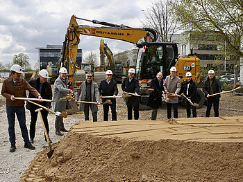 10 Männer in casual business outfit, 8 davon mit Bauhelmen, schippen beim Spatenstich mit Schaufeln Sand auf einer Baustelle, auf der ein Studierendenwohnheim entstehen soll. 