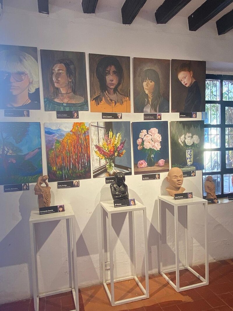 Blick auf 10 Bilder an einer Wand, davor 3 Skulpturen auf weißen Tischen. 