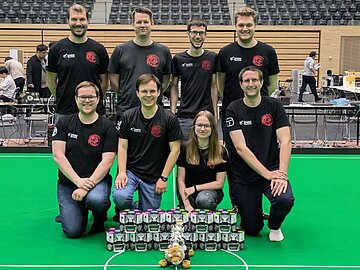 Gruppenbild mit 8 Personen auf grünem Roboterfußballfeld, davor 11 Fußballroboter und ein Tiger-Maskottchen.