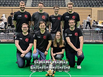 Gruppenbild mit 8 Personen auf grünem Roboterfußballfeld, davor 11 Fußballroboter und ein Tiger-Maskottchen.