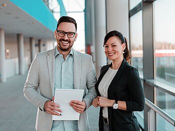 Zwei Personen im Business-Outfit, ein Mann links, eine Frau rechts. Beide lachen in die Kamera, er hält Zettel und Stift in der Hand.  