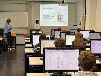Hörsaal von hinten zu sehen, auf den Tischen Monitore mit einem Simulationsprogramm, das Schüler*innen getestet haben. Vor der Gruppe steht ein Schüler, der zu einer Präsentation an der Wand etwas erläutert.
