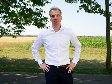Proträt Prof. Zippel in weißem Hemd und schwarzer Hose auf dem DHBW-Mannheim-Campus an einem sonnigen Tag mit Feld im Hintergrund.