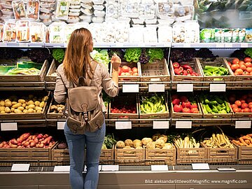 Eine Frau mit braunem Rucksack und Jeans steht von hinten her gesehen vor einem reich gefüllten Gemüseregal im Supermarkt und greift nach frischer Ware. Das Regal ist mit Holzkisten voller Tomaten, Paprika, Kartoffeln und Salat bestückt.