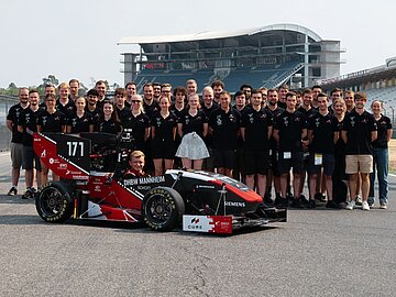 Teambild mit 40 Studierenden, davor der Rennwagen. Im Hintergrund eine Tribüne des Hockenheimrings. 