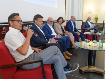 Sechs Menschen auf roten Samtstühlen auf einer Bühne sitzend, von der Seite fotografiert. Der erste davon spricht in ein Mikrofon.