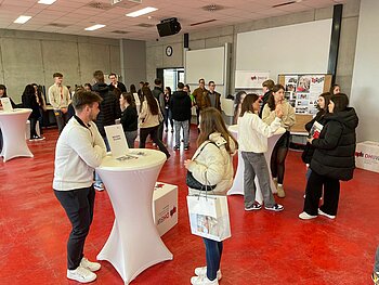 Raum mit vielen jungen Menschen an Stehtischen, die sich zu unterschiedlichen Studienrichtungen austauschen. 