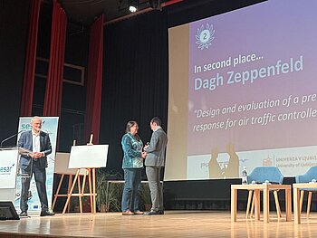 Blick auf eine Bühne aus dem Zuschauerraum rechts unten. Darauf sieht man einen Herrn ganz links an einem Pult, rechts daneben eine Dame, die Dagh Zeppenfeld den Award überreicht, rechts mehrere Stühle mit noch zwei jungen Erwachsenen, die der Überreichung zuschauen. Im Hintergrund auf großer Leinwand der Name von Hr. Zeppenfeld sowie der Titel seiner Bachelor-Arbeit.  