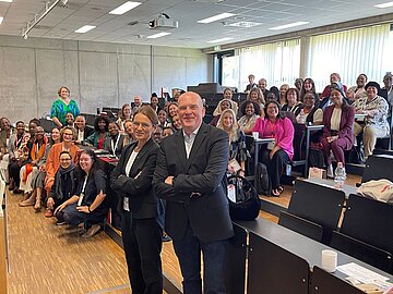 Im Hintergrund: Gruppe von ca. 40 Frauen im Hörsaal in Sitzreihen, davor ein Mann und eine Frau, die in die Kamera lachen. 