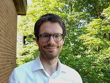 Portrait Prof. Dr. Maximilian Scherer neben einem Backsteinhaus und mit grünen Bäumen im Hintergrund.