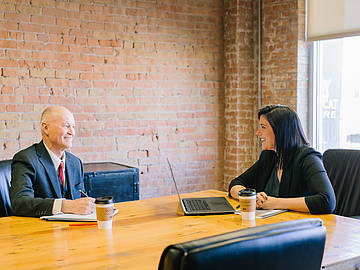Blick in ein Büro: Links sitzt ein älterer Herr, vermutlich kurz vor der Rente, im Anzug, etwas schreibend, er lächelt eine Frau Mitte 30 auf der anderen Seite des Tisches an, die vor einem Laptop sitzt und  zurücklächelt.
