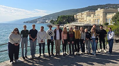 Gruppenbild mit 12 Studierenden am Hafen von Opatija mit Blick auf Meer und ein altes sandfarbenes Hotel an der Küste.