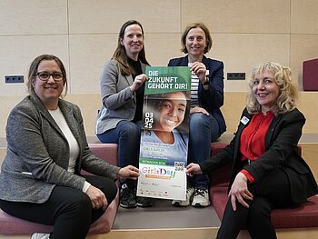 Gruppenbild mit 4 Frauenm die in die Kamera schauen und in der Mitte das Plakat zum Girls'Day halten. 