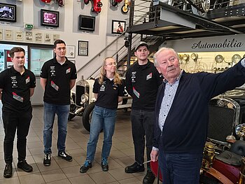 1 Studentin und 3 Studenten bekommen im Museum Autovision etwas von dem Besitzer erklärt. Er zeigt nach rechts im Bild, die Studenten folgen seinen Blicken. 