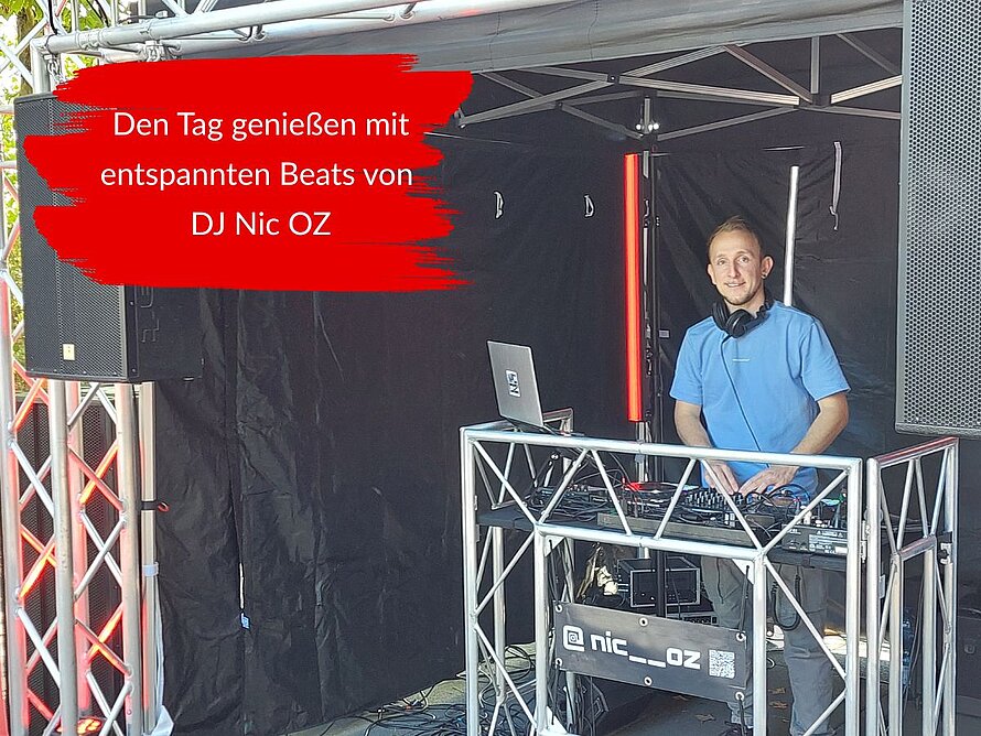 DJ Nic OZ steht beim Ersti-Welcome der DHBW Mannheim an seinem DJ-Pult mit Laptop und Mischpult. Um ihn herum stehen Lautsprecher und Veranstaltungstechnik. Der eingeblendete Text lautet: Den Tag genießen mit entspannten Beats von DJ Nic OZ.