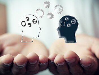 Zwei nach oben gerichtete Handflächen, darüber schweben zwei Grafiken von Köpfen im Profil, die zueinander schauen. Einer ist schwarz, der andere weiß, in beiden sowie darüber graue Zahnräder, die den Transfer von Wissen darstellen sollen. 