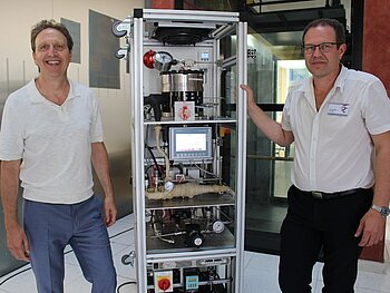 Prof. Dr. Wilhelm Wiebe und Prof. Dr. Volker Schulz posieren für ein Foto, sie stehen neben einem elektrochemischen Verdichter, der in einem Rollregal aufgebaut ist