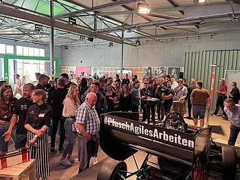 Menschenmenge in der U-Halle des Spinelli-Geländes. Im Hintergrund der Stand der DHBW Mannheim, vorn die Rückseite von Rennate auf deren Heckspoiler steht #Pfusch durchgestrichen und daneben Agiles Arbeiten.
