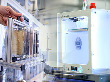 Collage aus zwei Bildern: Links hält eine Hand eine Halterung mit mehreren hintereinander gereihten Brennstoffzellen, rechts sieht man einen 3D-Drucker