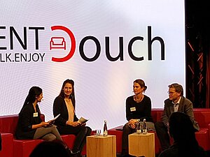 Vier Personen auf zwei roten Couches sitzen sich gegenüber und unterhalten sich, dahinter das Logo der Eventcouch