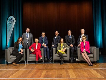 Gruppenbild mit 9 Personen auf einer Bühne, 4 in vorderer Reihe auf Sesseln sitzend, 5 in hinterer Reihe stehend.