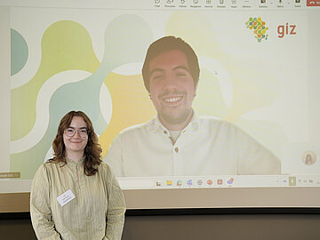 Im Vordergrund die Studentin Emilia Erasmus, im Hintergrund auf einer Präsentation der Student Christoph Eckinger, der online hinzugeschaltet wurde. Beide lachen freundlich in die Kamera. 