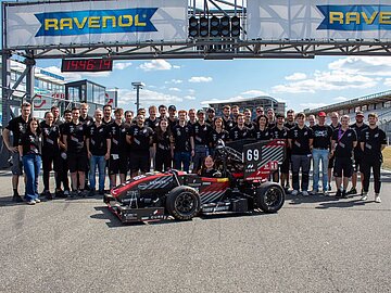 Gruppenfoto des Teams auf der Rennstrecke in der Mitte der Rennwagen mit der diesjährigen Fahrerin. 