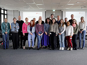 Gruppenbild mit 19 Personen in einem Besprechungsraum mit grauem Teppich und zwei Präsentationen im Hintergrund. 
