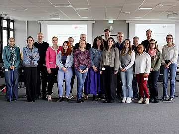 Gruppenbild mit 19 Personen in einem Besprechungsraum mit grauem Teppich und zwei Präsentationen im Hintergrund. 
