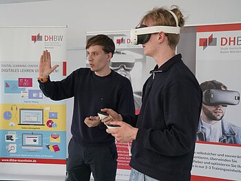 Zwei Männer in schwarzer Kleidung. Der rechte ist im Profil zu sehen, schaut nach links, hat eine VR-Brille an und dazugehörige Fernbedienungen in den Händen. Der linke macht eine erklärende Geste und schaut nach links.