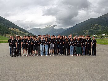 Gruppe von 42 Menschen in schwarzen CURE-T-Shirts auf einer Autorennstrecke vor einem schweizerischen Bergpanorama. 