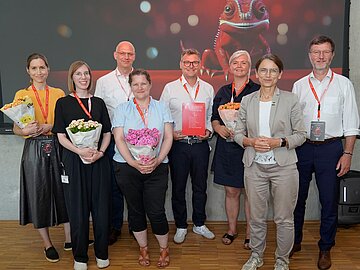 Gruppenbild mit 8 Personen, 4 davon mit Topfpflanzen in der Hand; dahinter das Keyvisual des diesjährigen Forschungstags: Camie, ein rotes Chamäleon. 