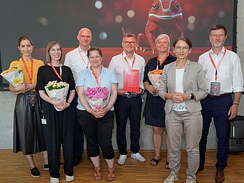 Gruppenbild mit 8 Personen, 4 davon mit Topfpflanzen in der Hand; dahinter das Keyvisual des diesjährigen Forschungstags: Camie, ein rotes Chamäleon. 