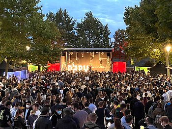 Menschenmenge mit Masken vor eine leuchtenden Bühne abends
