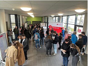 Blick von der Treppe ins Foyer mit Informationsstand und vielen Besucher*innen im Gespräch.